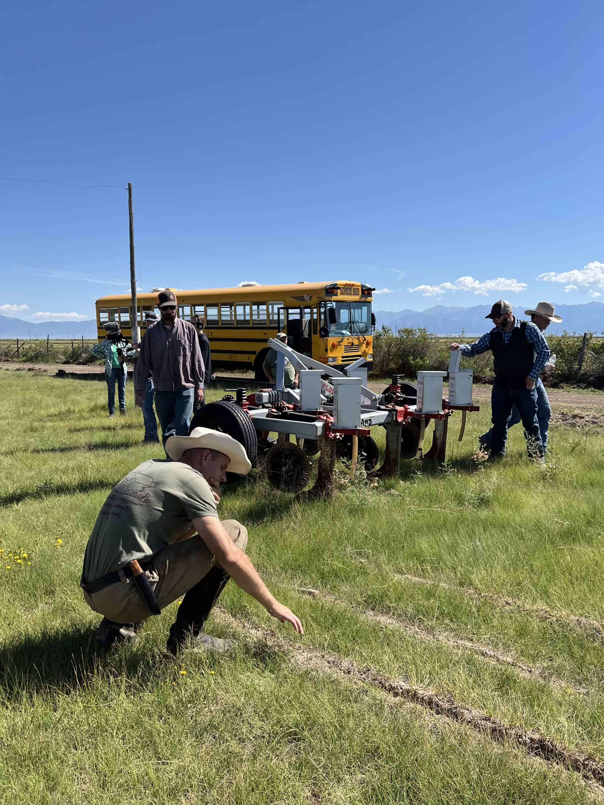 A man in a cowboy hat bends down to look at tillage rows in a green field while a group of people stand around a yeomans plow.